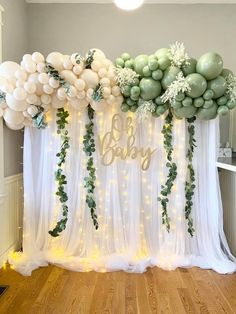 balloons and greenery decorate the backdrop for a baby shower with white sheer curtains, green garlands and lights