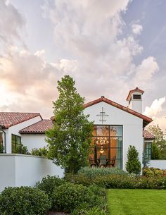 a white house with a cross on the roof and green bushes in front of it