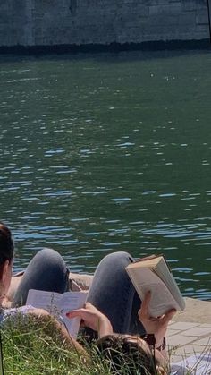 two people laying on the ground next to water reading books and looking at something in front of them