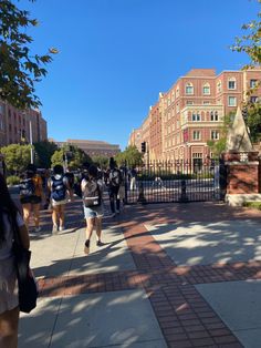 many people are walking down the sidewalk in front of some brick buildings and gated streets