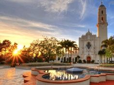 the sun is setting in front of a large building with a fountain and clock tower