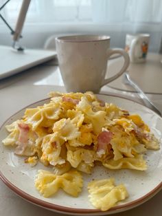 a white plate topped with pasta and ham next to a coffee cup on a table