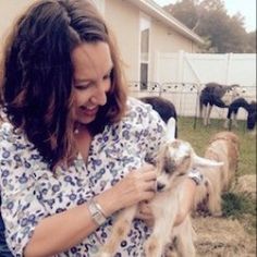 a woman holding a baby goat in front of other goats on the grass and fence