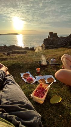 two people are sitting on the grass with food in front of them and water behind them