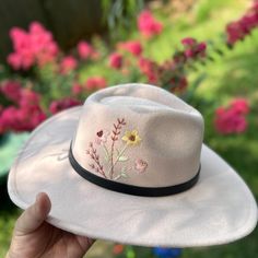 a hand holding a white hat with flowers on it in front of some pink flowers