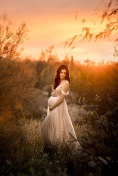 a pregnant woman in a white dress is standing in tall grass with the sun setting behind her