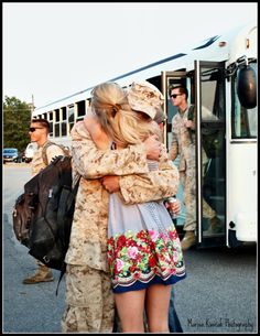 two people hugging each other in front of a white bus with soldiers standing behind them