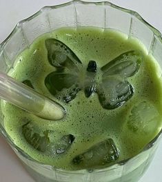 a pitcher filled with green liquid and ice cubes next to a plastic spoon on a white surface