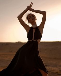 a woman in a black dress is dancing on the beach at sunset with her hands behind her head