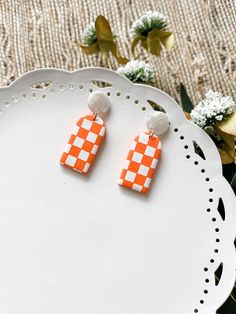 two orange and white checkered earrings sitting on top of a plate next to flowers