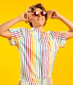 a woman with sunglasses on her head is posing in front of a yellow background wearing a colorful striped shirt