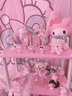 a pink table topped with lots of dolls and stuffed animals next to a hello kitty wall