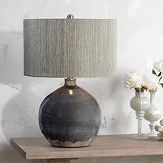 a gray vase sitting on top of a wooden table next to a lamp and flowers