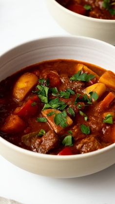 two bowls filled with stew and vegetables on top of a white tablecloth next to each other