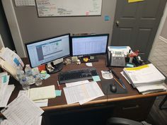 two computer monitors sitting on top of a desk covered in papers and other office supplies