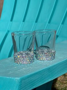 two shot glasses sitting on top of a blue bench next to each other with glitter in them