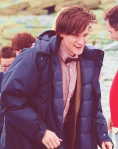 a man in a coat and bow tie is standing on the beach with other people