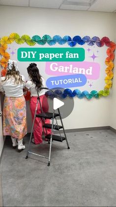 two women standing in front of a sign that reads diy paper fan garland garland