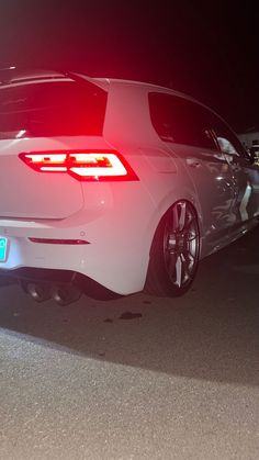 the back end of a white car parked in a parking lot at night with its lights on