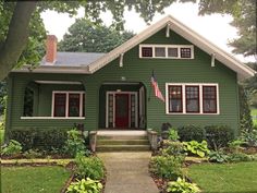 a green house with white trim and red windows on the front door is surrounded by greenery