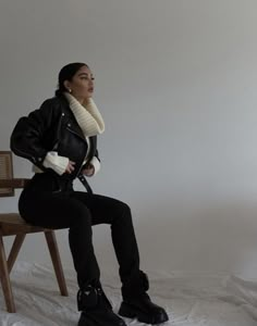 a woman sitting on top of a wooden chair next to a white wall and wearing black boots