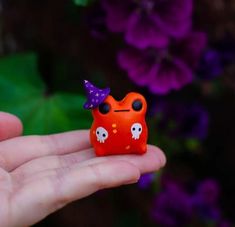 a hand holding an orange toy with two skulls on it and a purple flower in the background