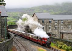 a train traveling down tracks next to a stone building and green hills in the background