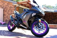 a black motorcycle with purple rims parked in front of a brick building on a sunny day