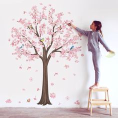 a woman is painting a tree with pink flowers on the wall in front of her