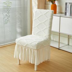 a white chair sitting in front of a window covered in ruffles and a vase with flowers