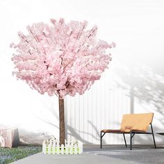 a tree with pink flowers in front of a white fence and wooden chair next to it