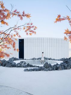 a person standing in front of a white building surrounded by rocks and trees with red leaves