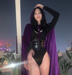 a woman in a purple cape and black bodysuit posing for the camera with her hands on her head