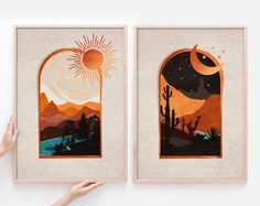 two framed art pieces depicting desert scenes with sun and moon in the sky, one being held up by someone's hand