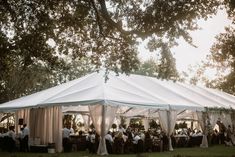 a group of people sitting at tables under a white tent in the middle of a park