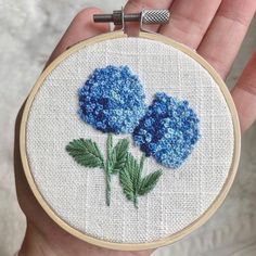 a hand holding a blue flower embroidered onto a white hoop