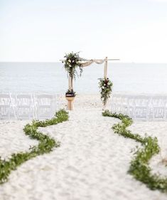 an outdoor ceremony set up on the beach with chairs and greenery draped over it