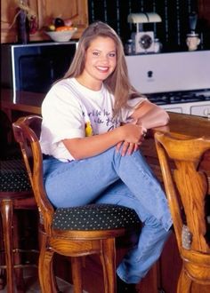 a young woman sitting on top of a wooden chair next to a kitchen counter with an oven in the background