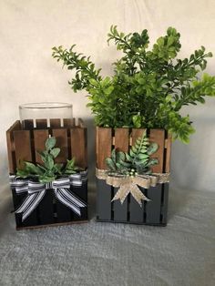 two wooden crates with plants in them on a table next to a glass vase filled with greenery