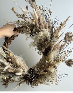 a wreath made out of dried plants and pine cones is being held by someone's hand