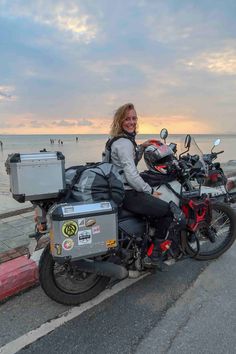 a woman sitting on a motorcycle next to the ocean with her luggage strapped to it