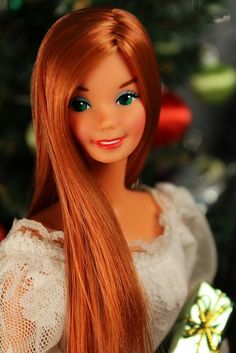 a doll with long red hair and green eyes holding a present in front of a christmas tree