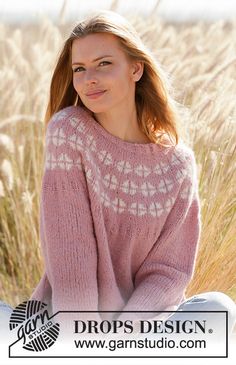 a woman sitting on the ground in front of some tall grass wearing a pink sweater