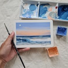 a person holding a paintbrush next to some watercolors and a painting on a table