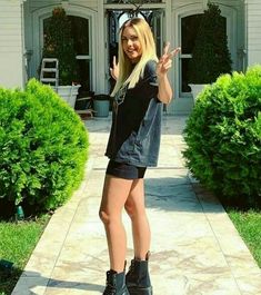 a woman standing in front of a house with her hand up to the camera and wearing black boots