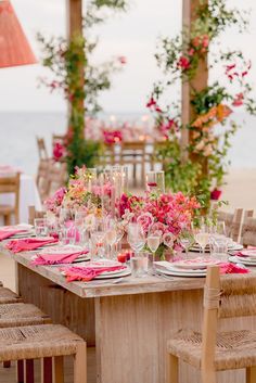 the table is set with pink flowers and wine glasses for an outdoor wedding reception on the beach