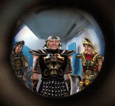 two men dressed in armor are seen through a magnifier lens as another man looks on