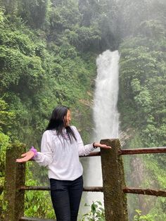 a woman standing next to a wooden fence in front of a waterfall with her arms outstretched