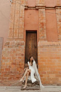 a woman sitting in front of a brick building wearing a white dress and holding a veil