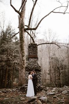 a bride and groom standing in front of an old stone fire place surrounded by trees